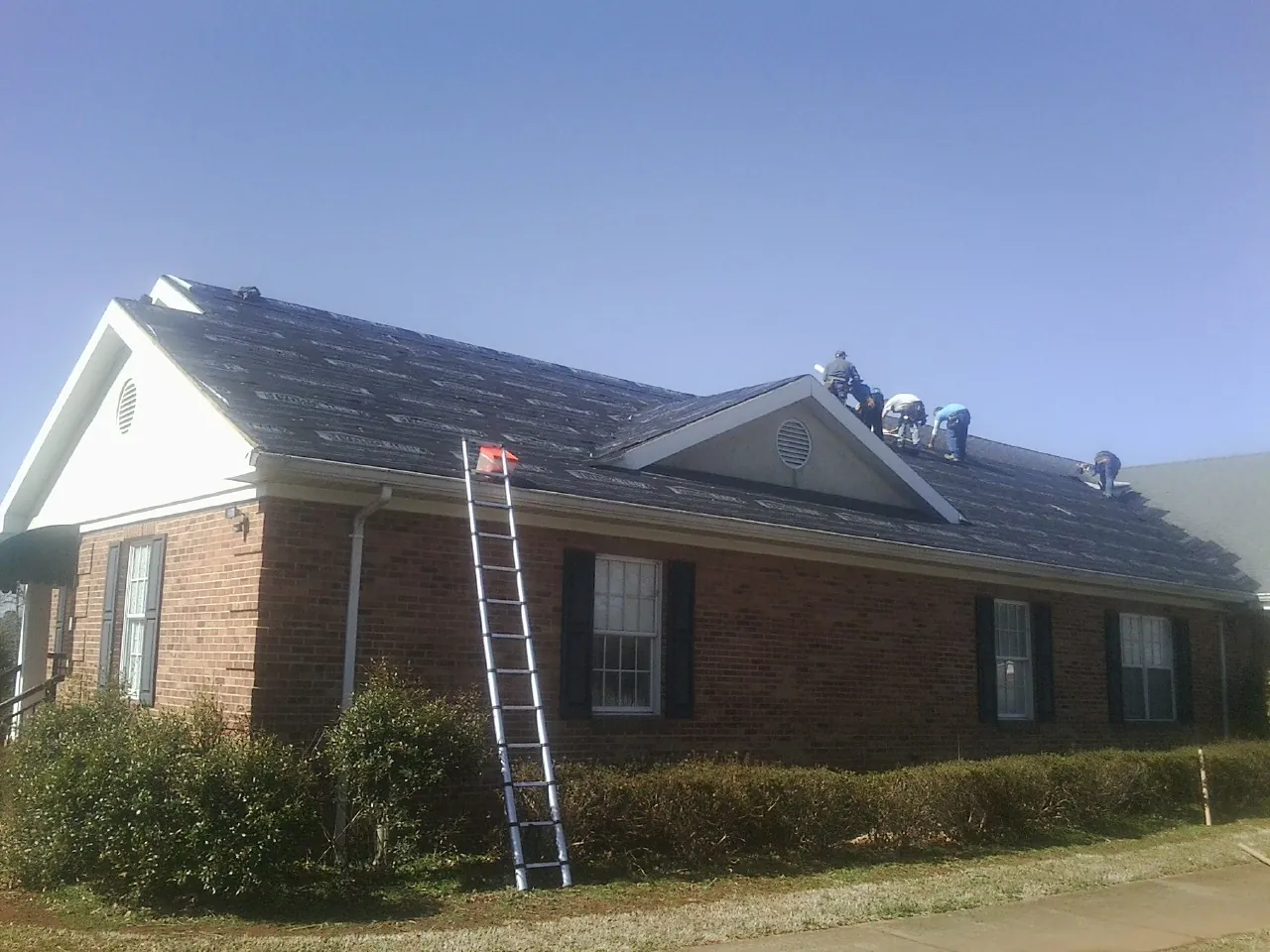 Crew tearing shingles off a brick ranch roof with a ladder set — Emergency Roof Repair work by our crew in Remsenburg