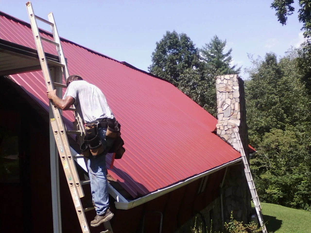 Roofer on a ladder beside a red metal roof and stone chimney — Soffit & Fascia Repair by Roofing Brothers in Remsenburg